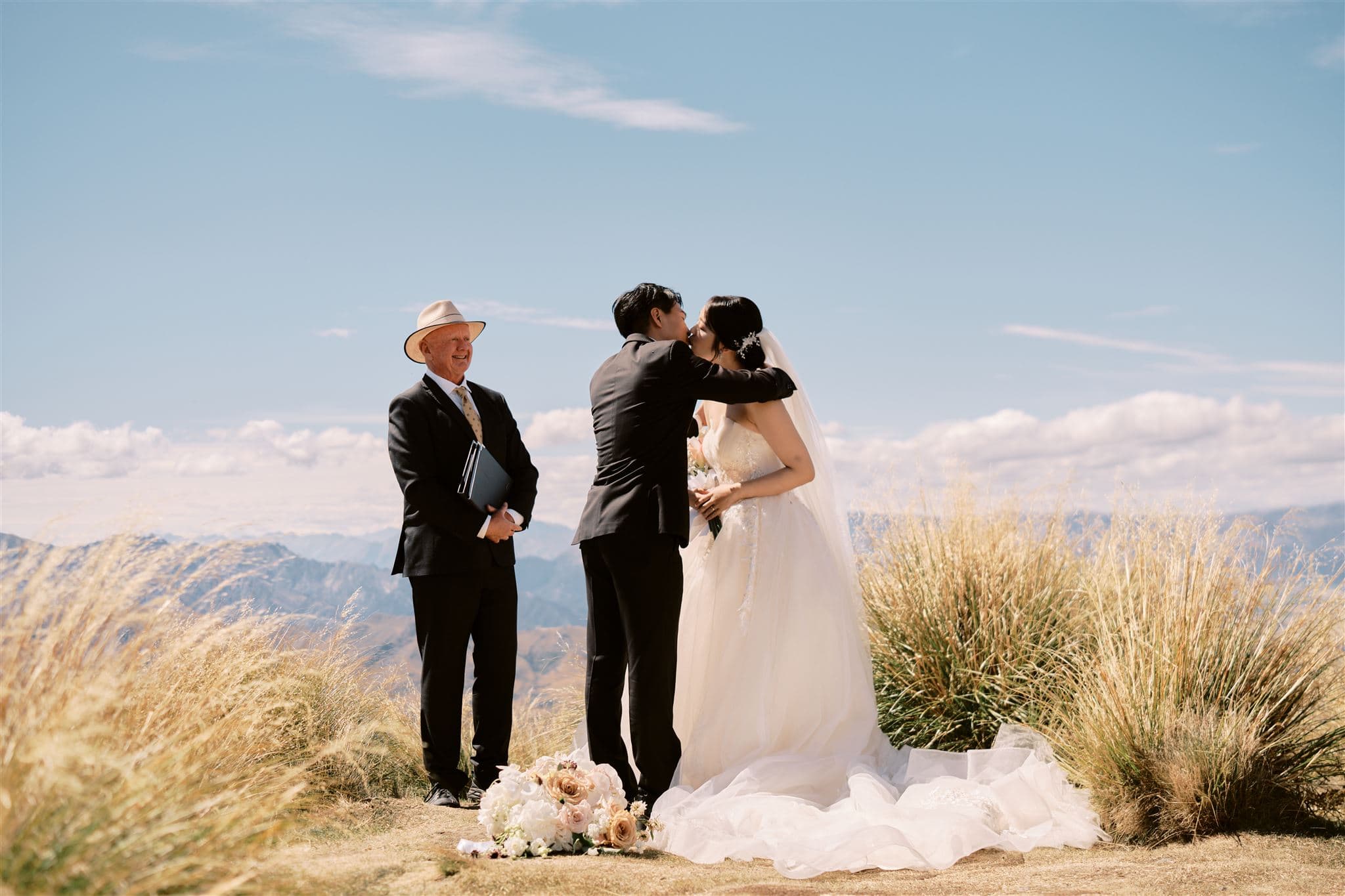 Queenstown New Zealand Heli Wedding Elopement Photographer クイーンズタウン　ニュージーランド　エロープメント 結婚式 | A couple exchanging a kiss at their outdoor wedding ceremony in Queenstown, officiated by a man in a hat, with scenic mountains in the background.